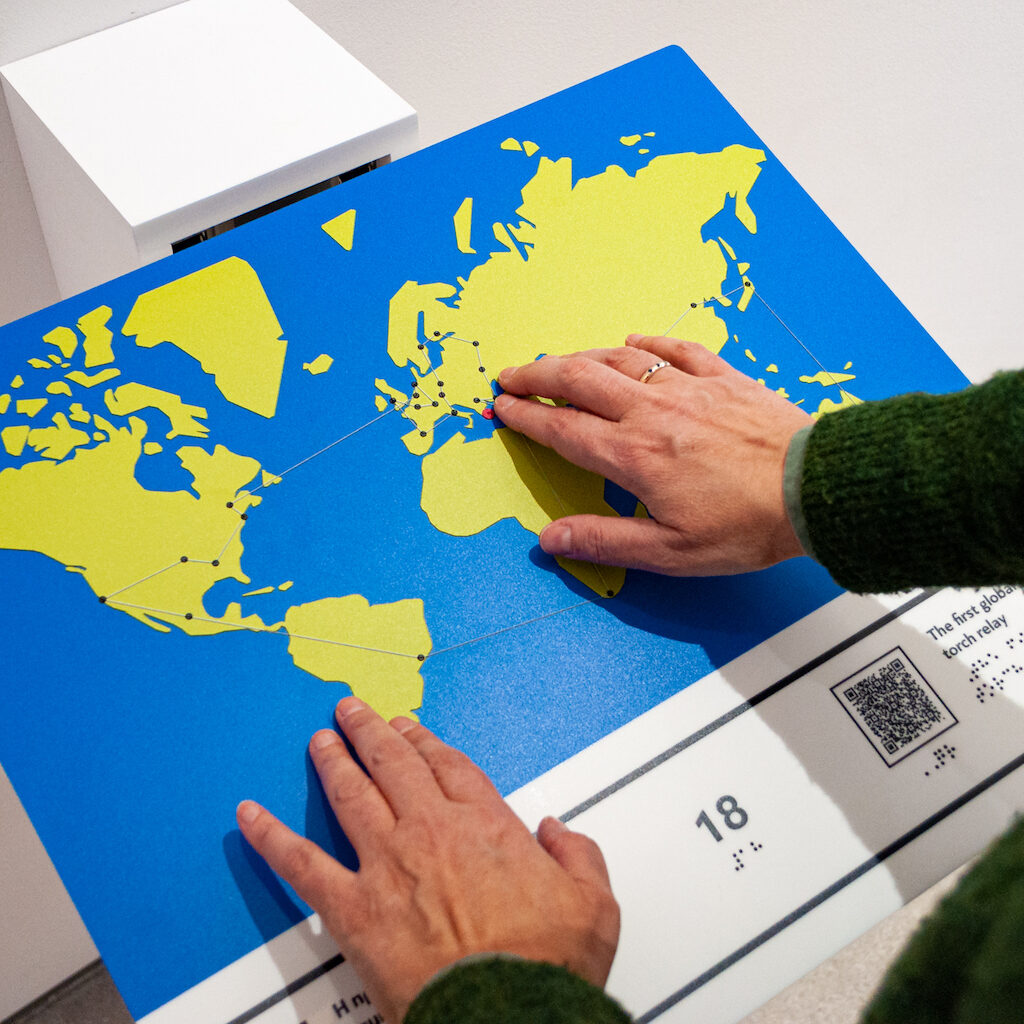 Tactile Representation of the First Global Torch Relay Performance held in the Olympic Games of Athens 2004, which is part of the Listen to Our Story Project of the Athens Olympic Museum. On the image appear a pair of hands exploring the tactile route on the relief world map. The Land is in light green color against the blue back ground of the oceans. Lower appears the number 18 in large font and in braille transcription along with a QR code.