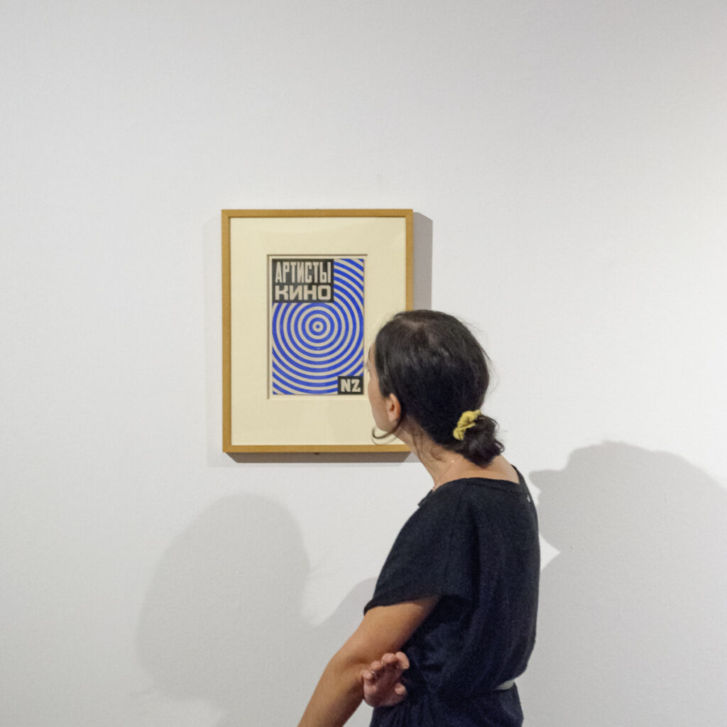 A woman with black hair and a black blouse stands in front of the artwork hanging on the wall and observes it carefully.