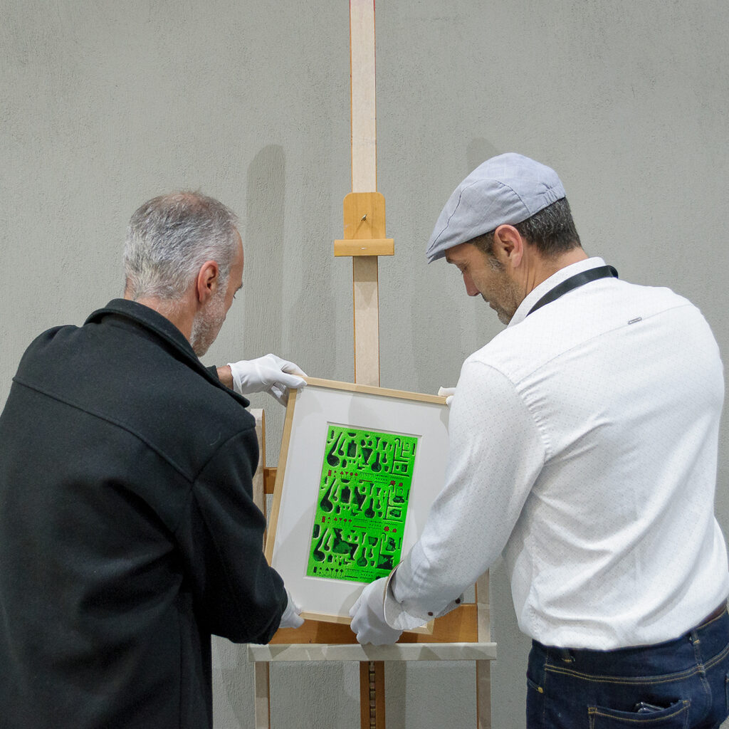 Two men wearing white gloves carefully place the artwork Alchemy Β on the easel.