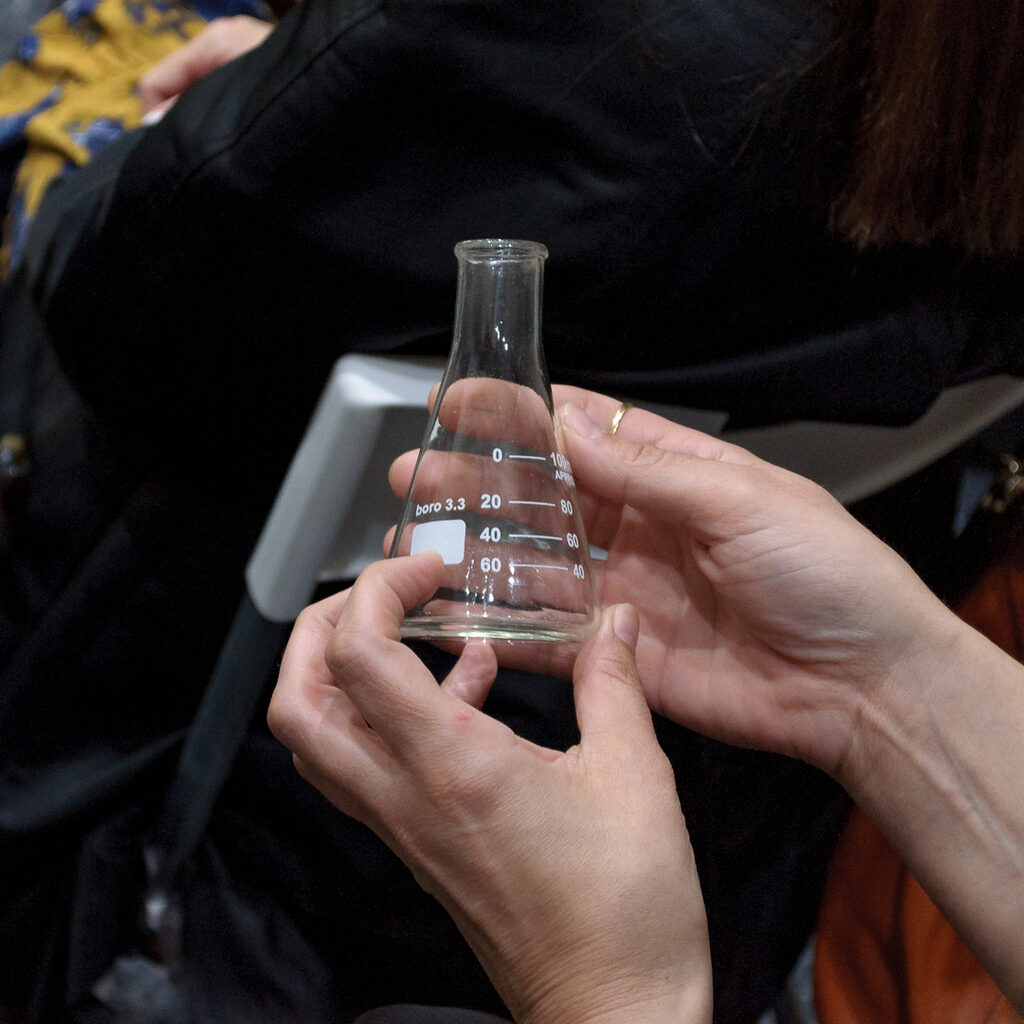 A pair of hands are prying into a small glass container, a conical volumetric flask with a narrow mouth.