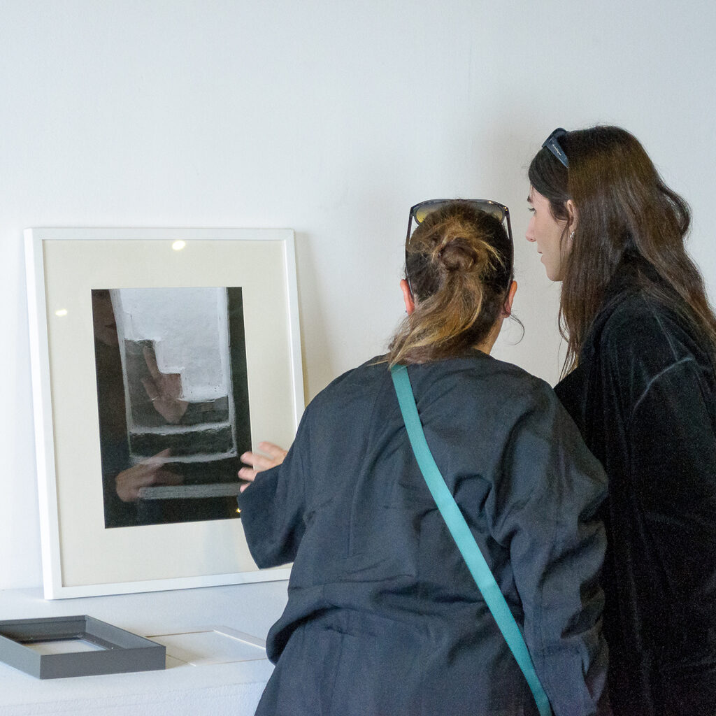 A black and white photograph rests on a white table leaning against a white wall. Two women stand in front of it and observe it.