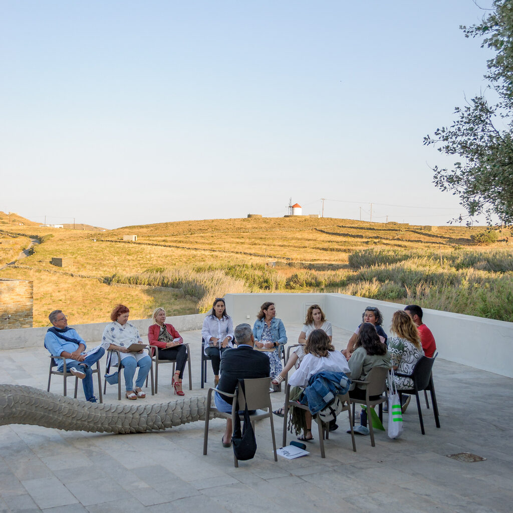 Audience participating in the Meeting Point 04 sits outside around a sculpture from which the tip of a snake's tail can be seen. In the background, the landscape of Tinos is visible in the afternoon sun.