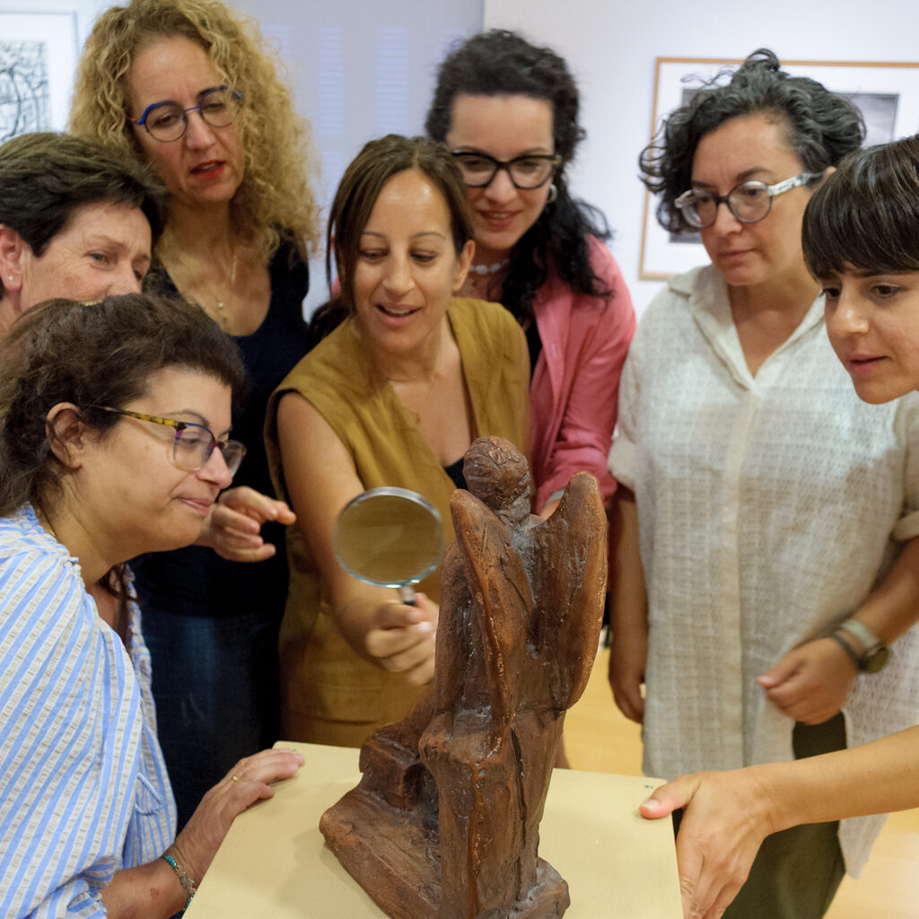 Κοινό έχει συγκεντρωθεί γύρω από ένα μικρό άγαλμα πάνω σε βάθρο. Μια γυναίκα στο κέντρο κρατάει στο χέρι της ένα μεγεθυντικό φακό.