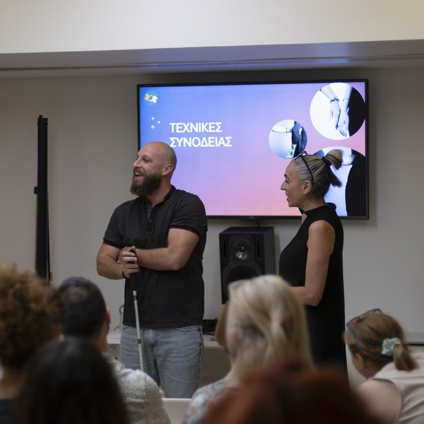 Άνδρας με λευκό μπαστούνι και γυναίκα μπροστά από οθόνη με παρουσίαση.