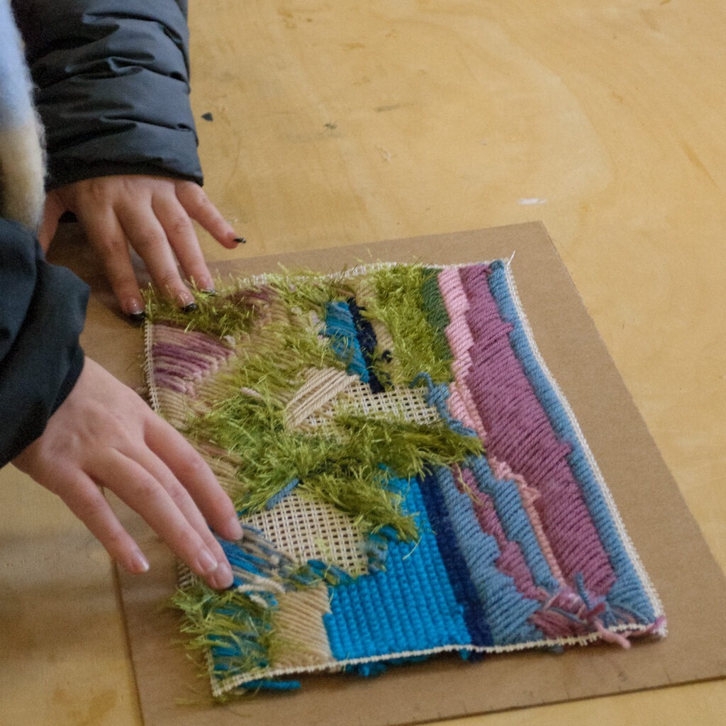 Χέρια αγγίζουν ένα πλεκτό έργο τέχνης πάνω σε ξύλινη επιφάνεια.