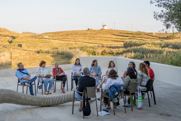 A group of people seated in a circle takes part in a discussion in an outdoor setting, with an agricultural landscape and low hills in the background.