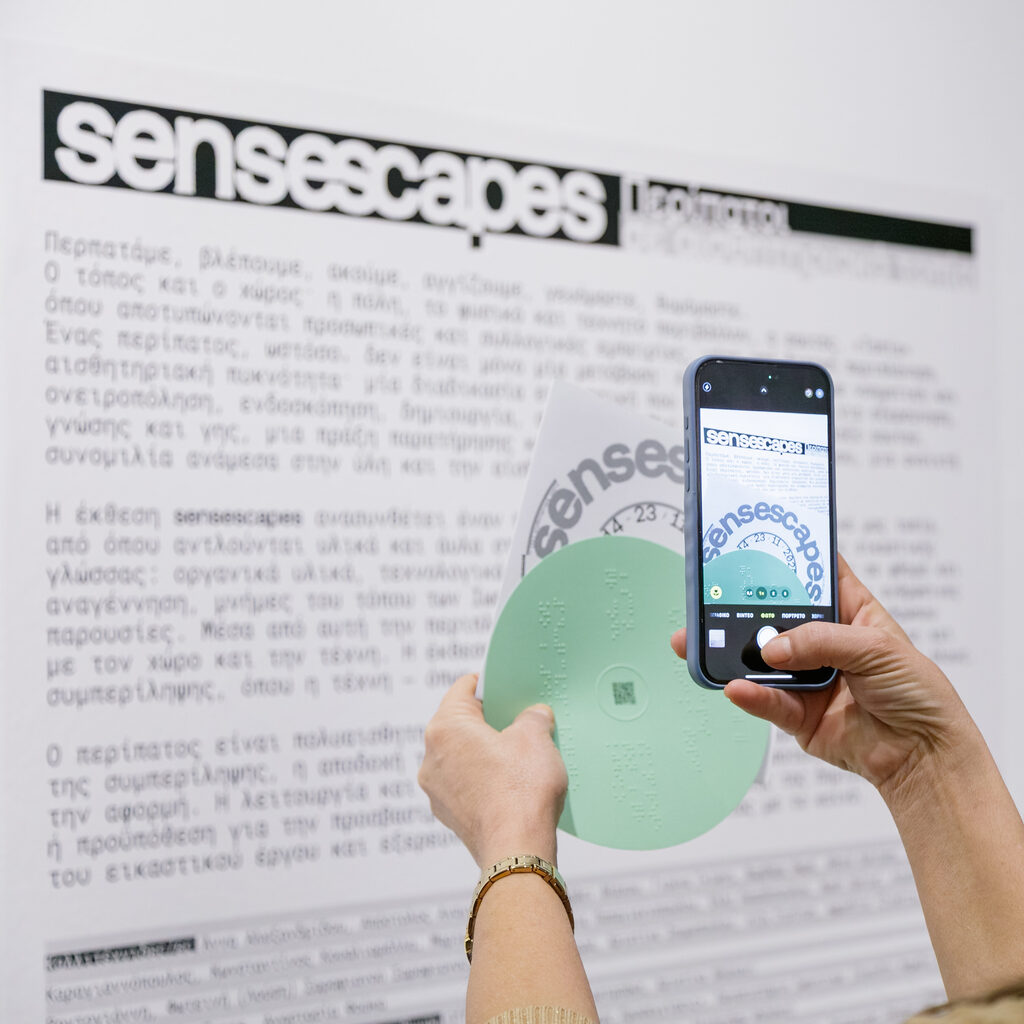 A person holds a mobile phone in one hand taking a picture of the circular exhibition flyer that holds with the other hand in front of the exhibition information panel titled “sensescapes”.