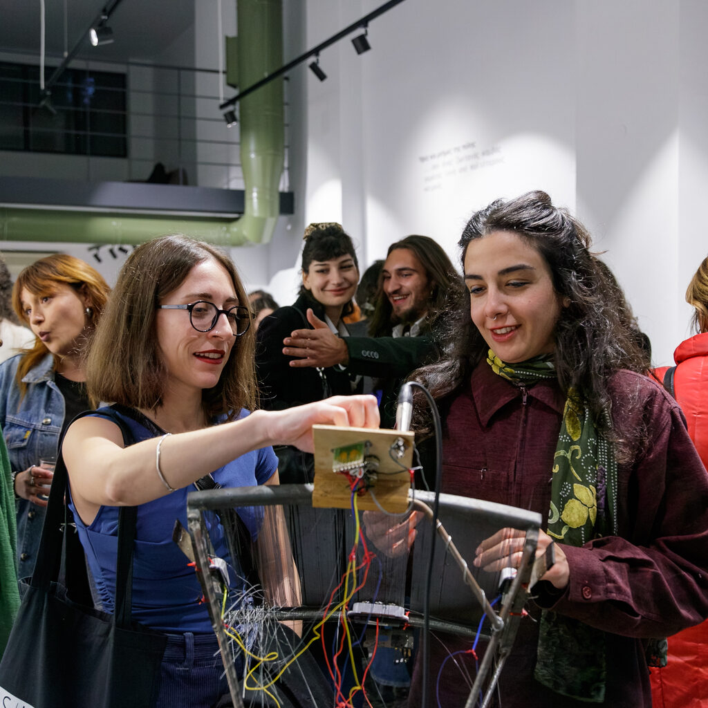 In the crowded exhibition space, two visitors interact with an electronic installation featuring wires and electronic components, as others watch smiling.