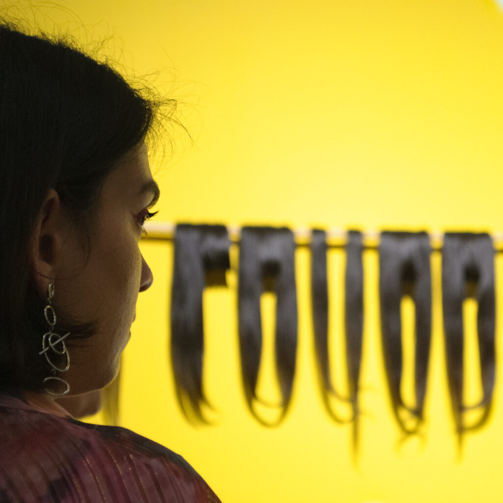Woman observing installation with black locks of hair in front of yellow wall