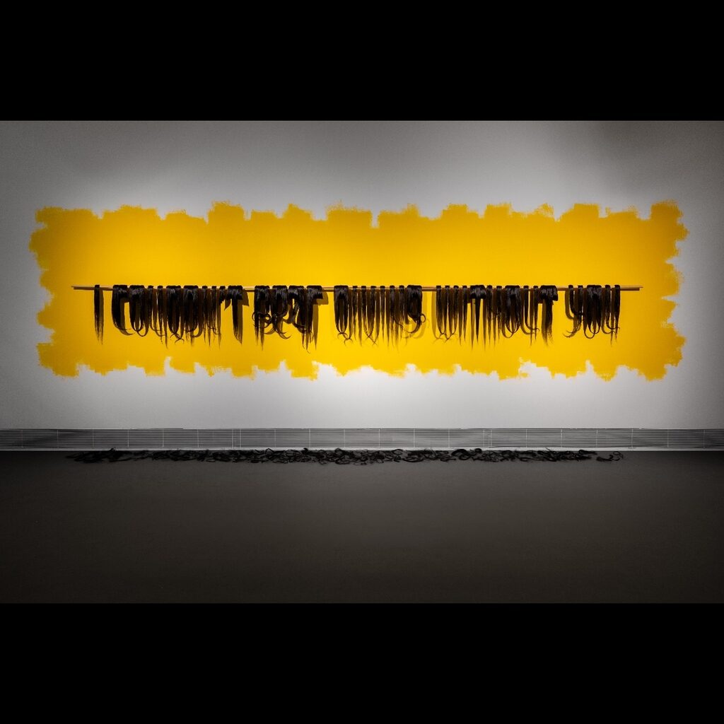 Installation with black hair strands hanging from a rod against a yellow background.