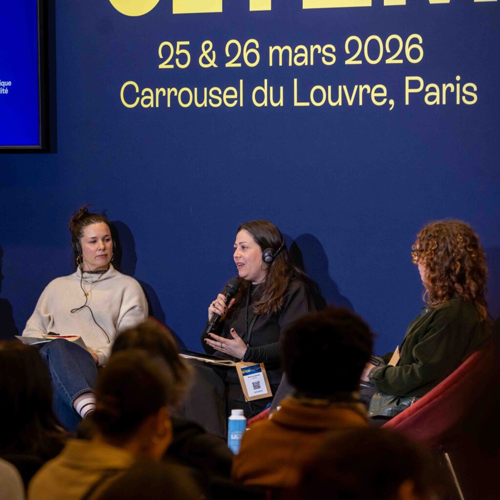 Three of the four panel speakers are in discussion on stage. Stela Anastasaki speaks while wearing translation headphones, as the others listen attentively.
