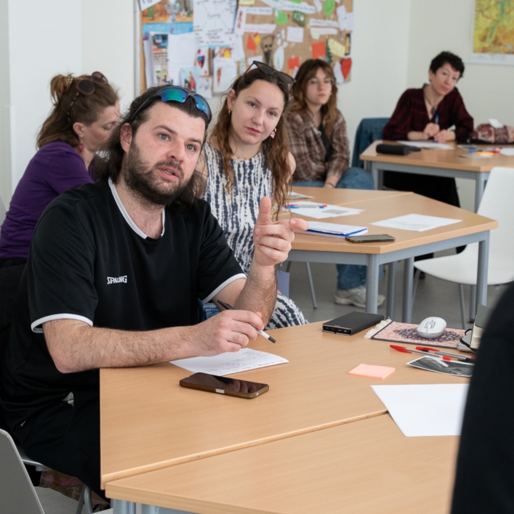 A participant speaks while other participants in the background are listening and taking notes during a workshop.
